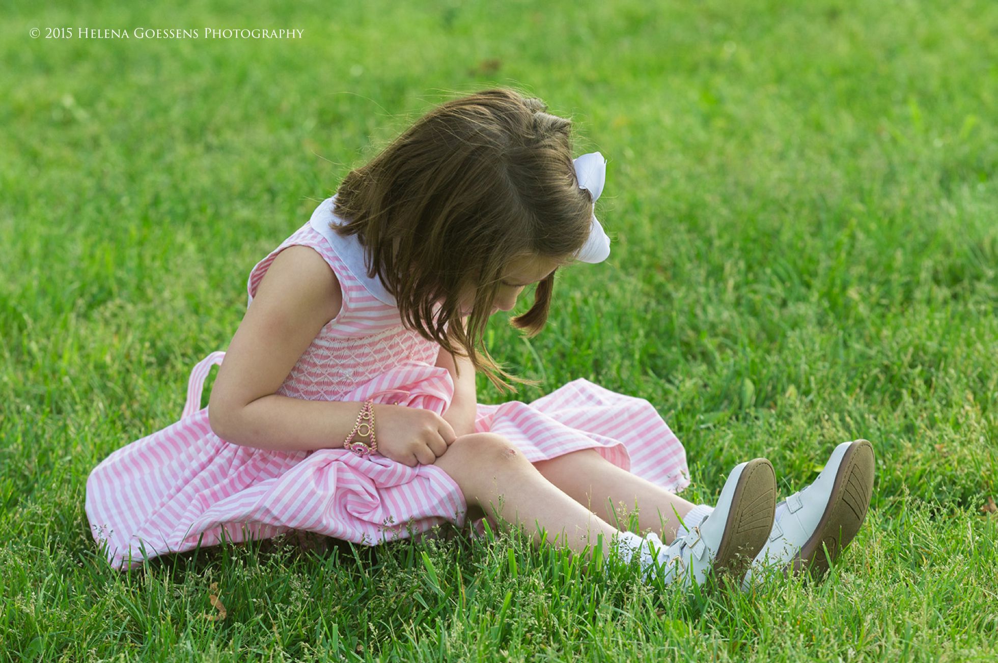 little girl with pink dress looking at her knee, sitting on the green grass
