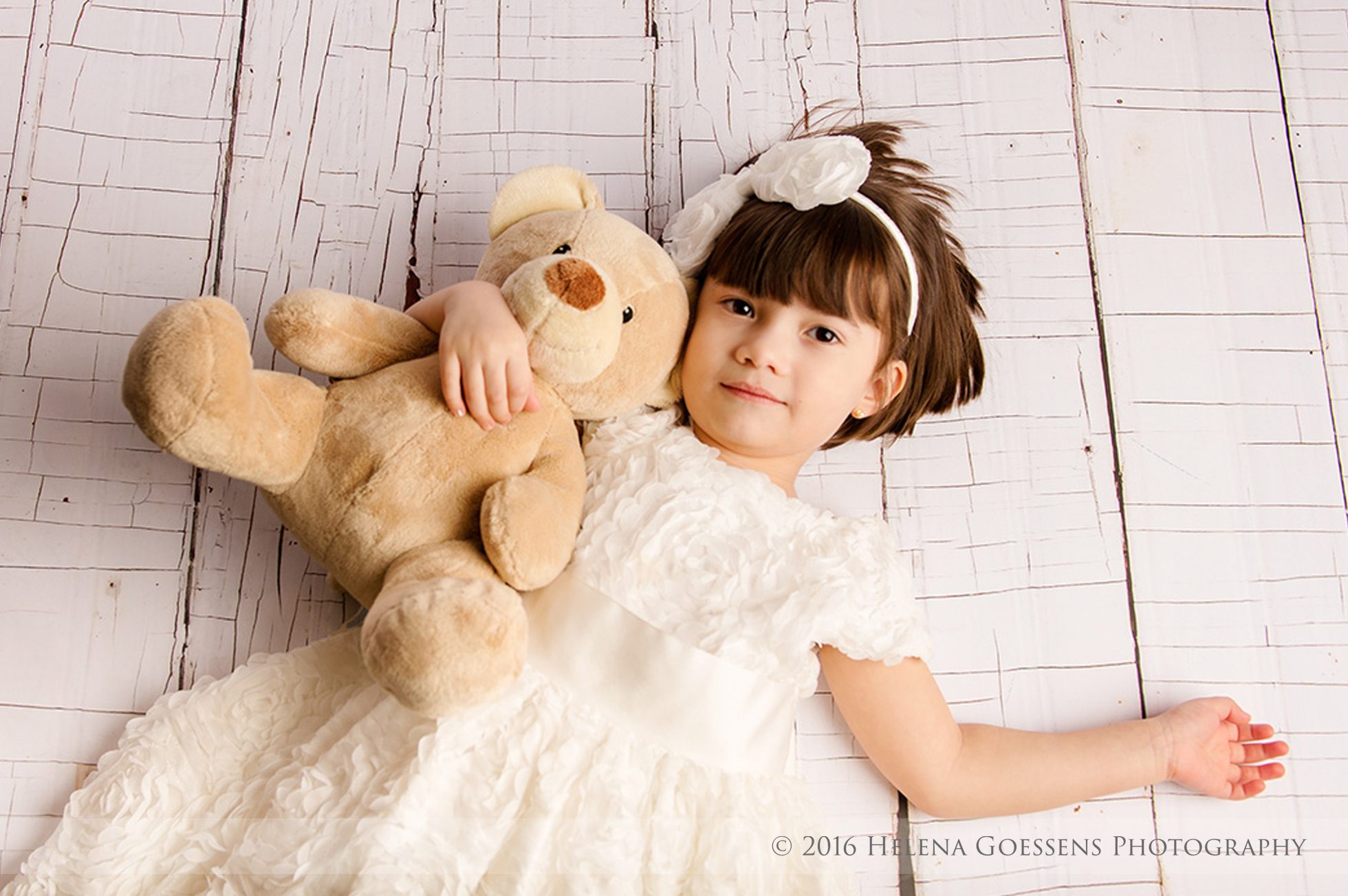 little girl in a white dress and a white bow tie holding her cream color teddy bear