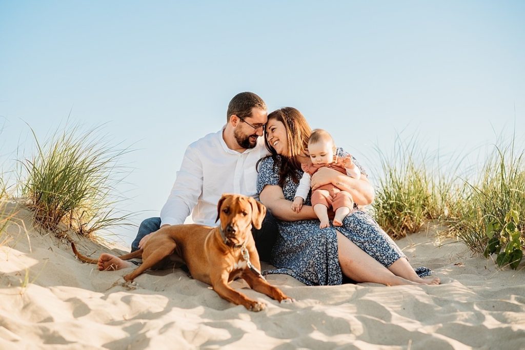 family sits on beach holding toddler and dog during summer photo sessions 