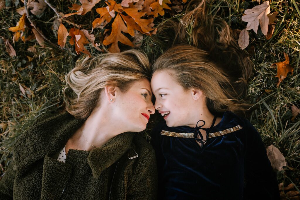 mother and daughter lay on grass with orange leaves in brown jackets during fall family portraits in Boston MA 