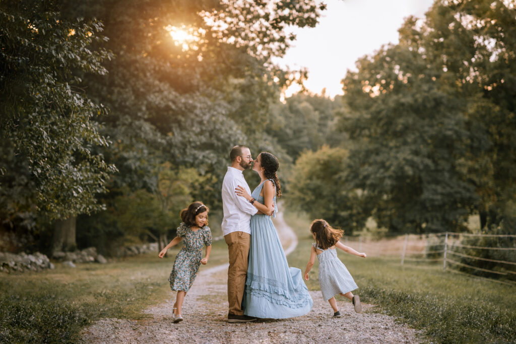 parents kiss on path while two girls run around them during Spring Family Photos with Helena Goessens Photography