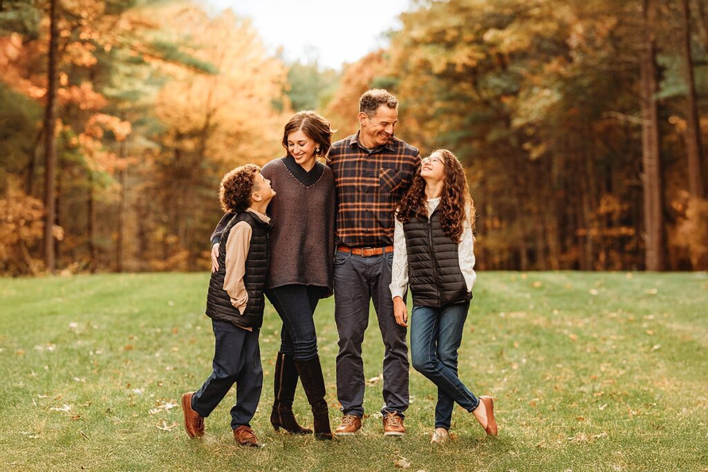 A happy family of four stands together on a grassy field with colorful autumn trees in the background, smiling at each other. This scene beautifully captures how families cherish special moments outdoors.