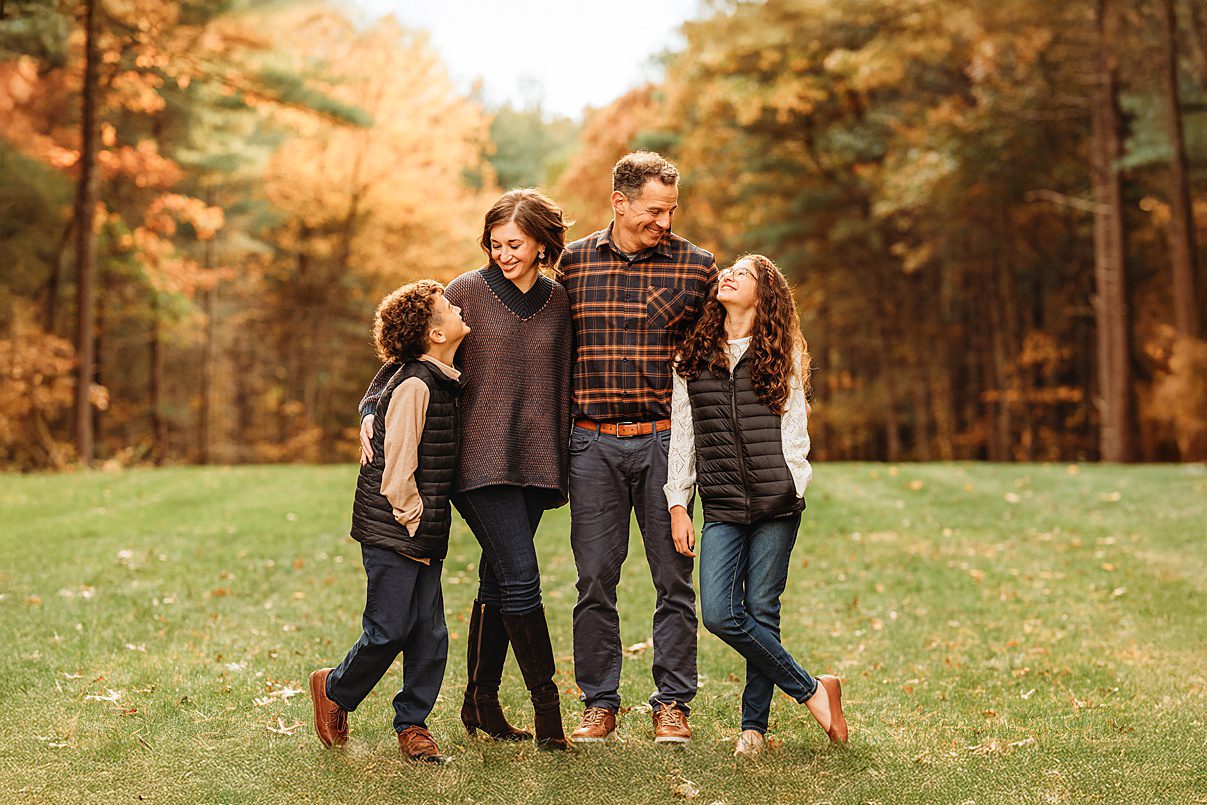 A happy family of four stands together on a grassy field with colorful autumn trees in the background, smiling at each other. This scene beautifully captures how families cherish special moments outdoors.
