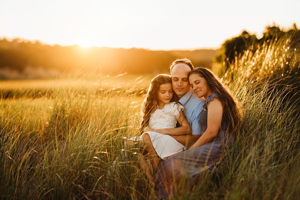 A family of three sits closely together in tall grass at sunset, with warm sunlight glowing around them. Like many families, the parents and their young daughter have their eyes closed, appearing peaceful and content.