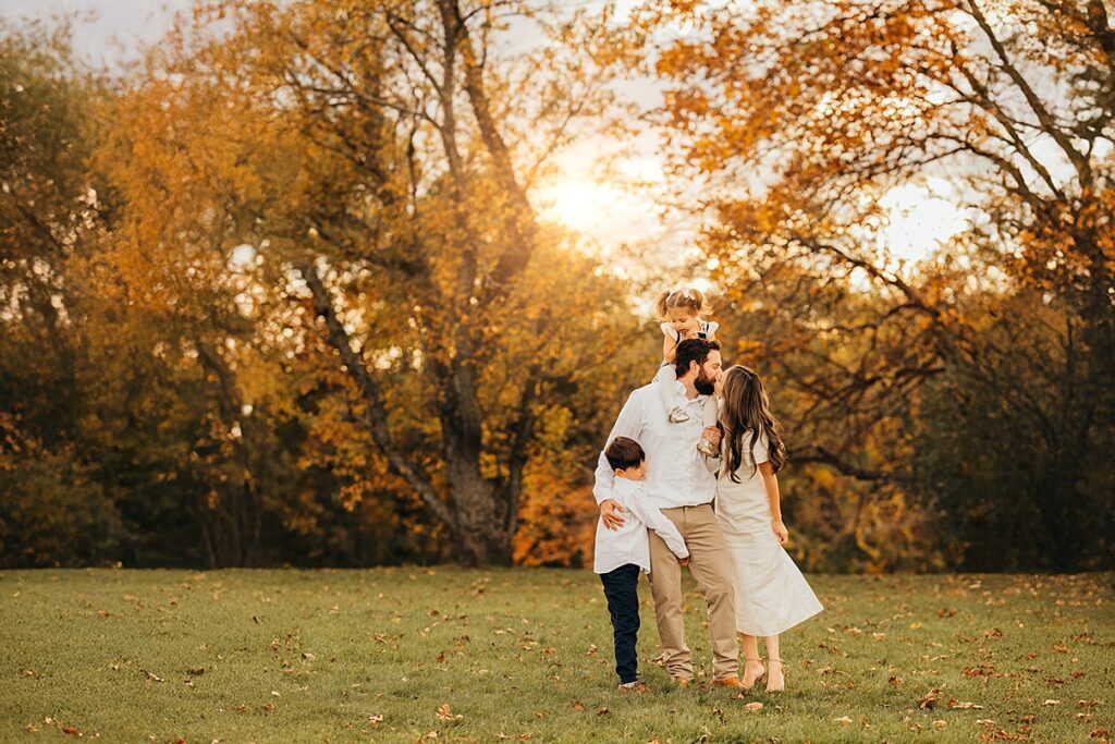 A family of four stands together on a grassy field with autumn trees in the background. The parents kiss while their two children, dressed in white, hug them—capturing the warmth and love that families share under the golden setting sun.