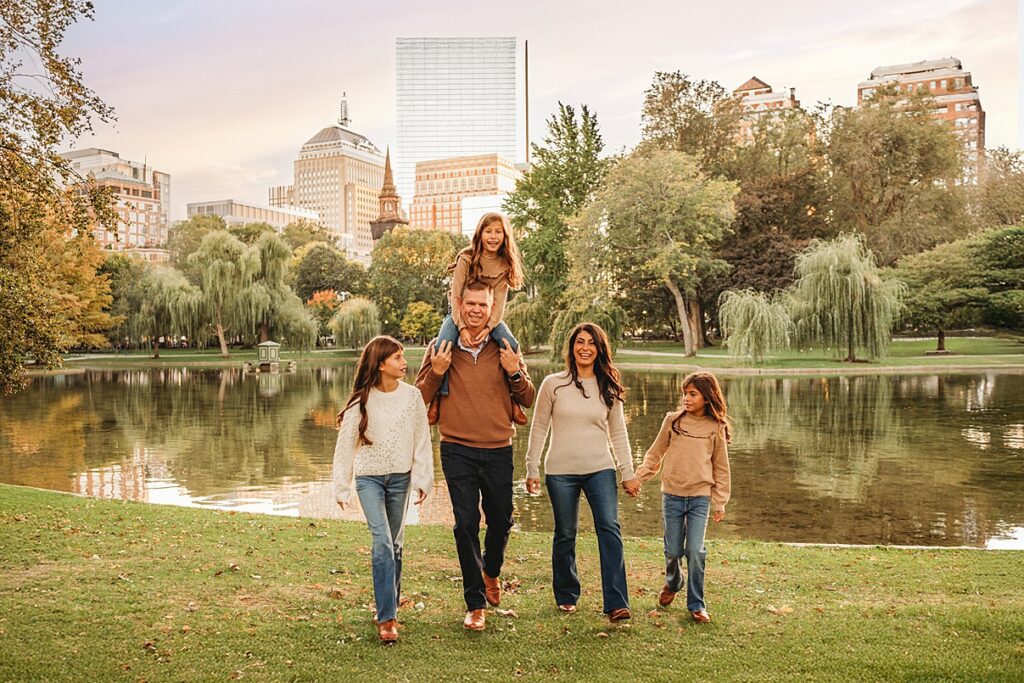 A family of five walks together by a pond in a park, with two adults and three children. Like many families enjoying time outdoors, the father carries one daughter on his shoulders. Trees and city buildings are visible in the sunset background.