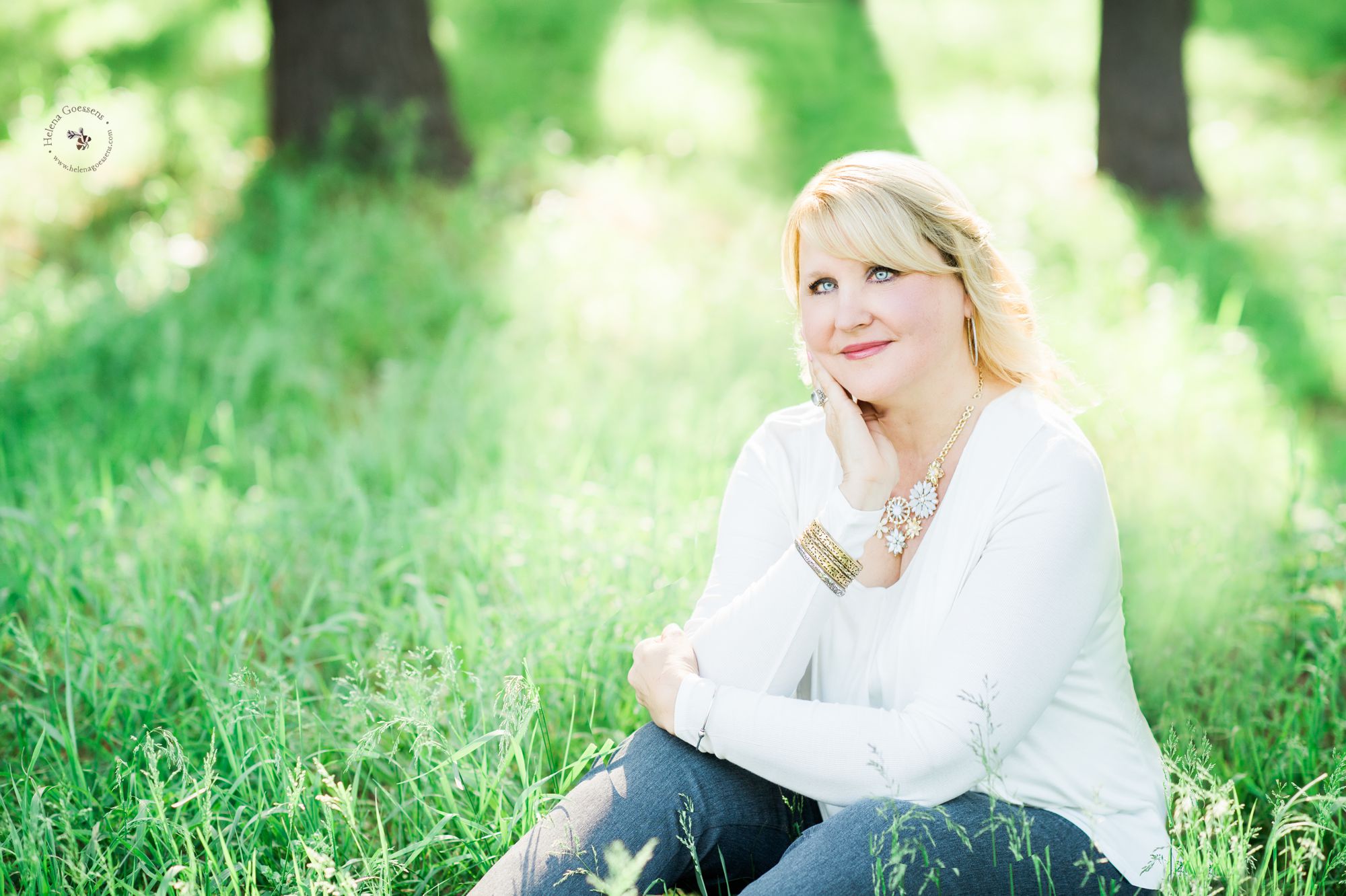 blond woman with blue eyes wearing a white top sitting on the grass