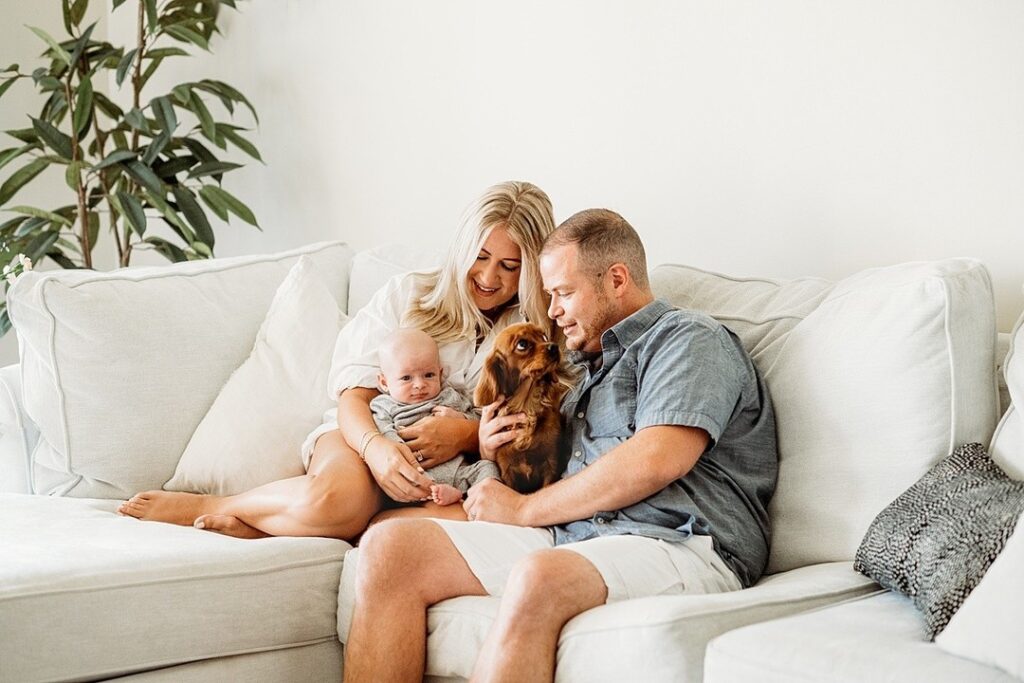 A couple sits on a white coach with their baby and a small dog, captured beautifully by Helena Goessens Photography. The man a tthe dog while the woman embraces the baby. The dog looks lovingly towards the man, completing this touching family scene.
