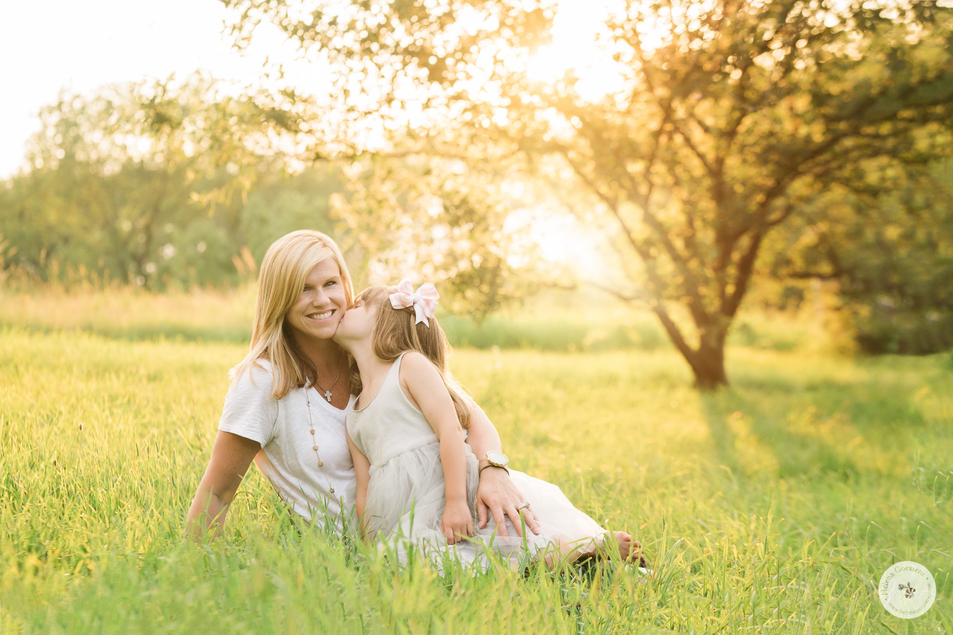 Daughter kissing mom. Both sitting on the grass at sunset. Image by Helena Goessens Photography.