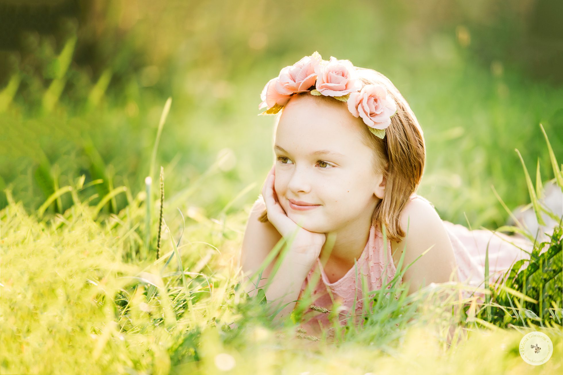 Portrait of a girl in a pink dress lying on the grass and wearing a pink roses headband for a Family Summer Session. Image by Helena Goessens Photography.