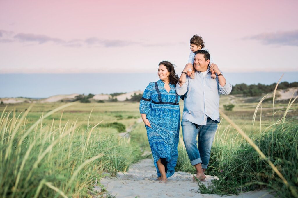 A family of three walks barefoot on a sandy path through grassy dunes at Rexhame Beach. The child sits on the father’s shoulders, while the mother walks beside them, all smiling beneath a pastel pink and blue sky—perfect for family portraits.