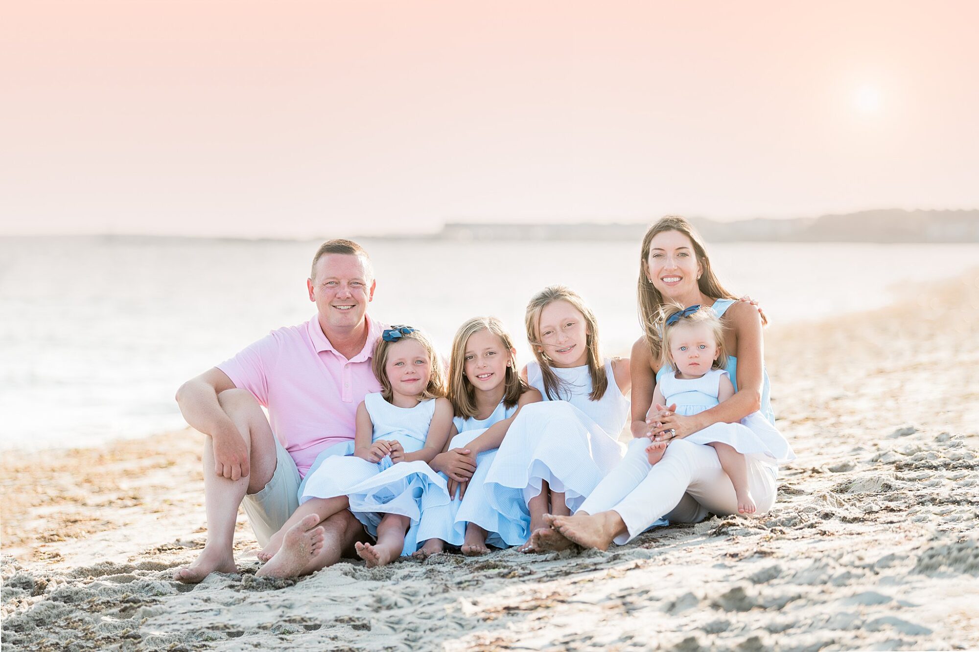 A family of six sits on a sandy beach at sunset for their Rexhame Beach Family Portraits. The parents, two girls, and two younger children wear pastel clothing, smiling together with the ocean and soft pink sky in the background.