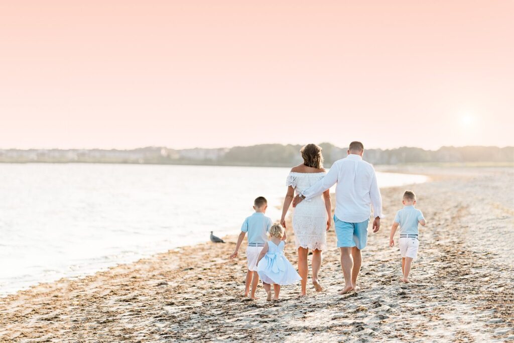 A family of five, dressed in light blue and white, walks hand in hand along a sandy shore at sunset. The sky glows pink as the sun dips low—an ideal scene for timeless Rexhame Beach Family Portraits.