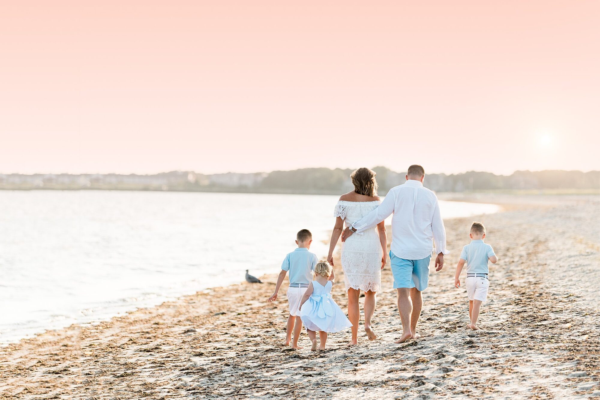 A family of five, dressed in light blue and white, walks hand in hand along a sandy shore at sunset. The sky glows pink as the sun dips low—an ideal scene for timeless Rexhame Beach Family Portraits.