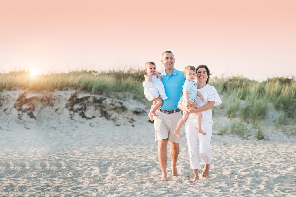 A family of four walks barefoot on Rexhame Beach at sunset. The parents each carry a young child and smile, with dune grass and a pastel pink sky in the background—perfect for memorable Rexhame Beach family portraits.