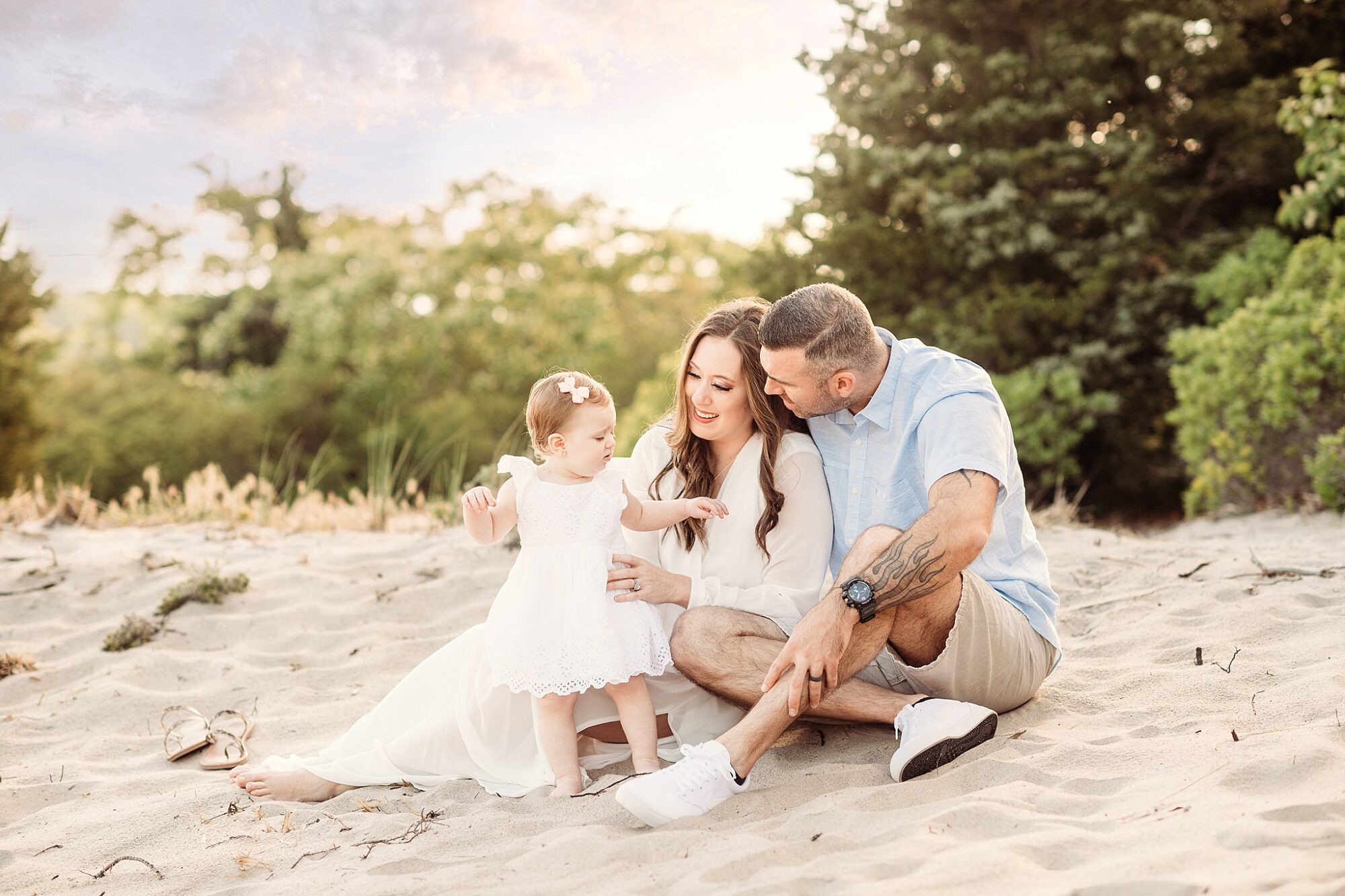 A young family sits on sand surrounded by greenery in this bright and serene Rexhame Beach family portraits session. The mother and father smile lovingly at their toddler daughter, who stands between them in a white dress.