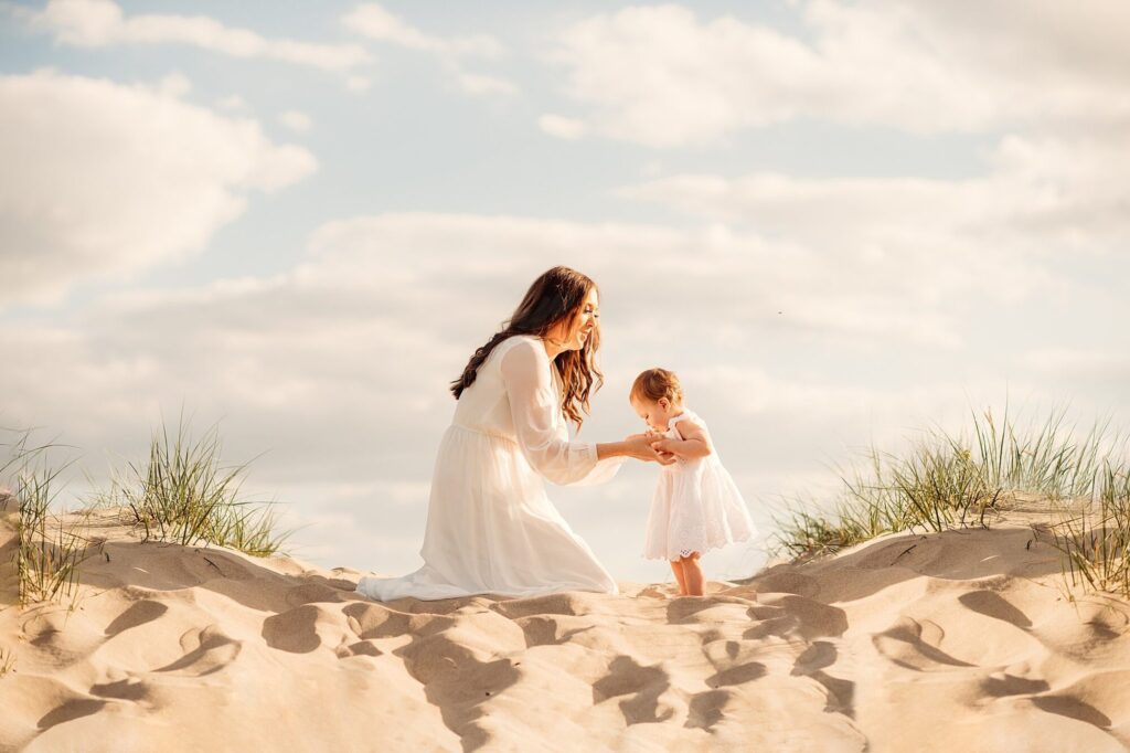 A woman in a white dress kneels on sandy dunes at Rexhame Beach, holding hands with a small child. Surrounded by tall grass beneath a bright, partly cloudy sky, their moment captures the essence of family portraits.