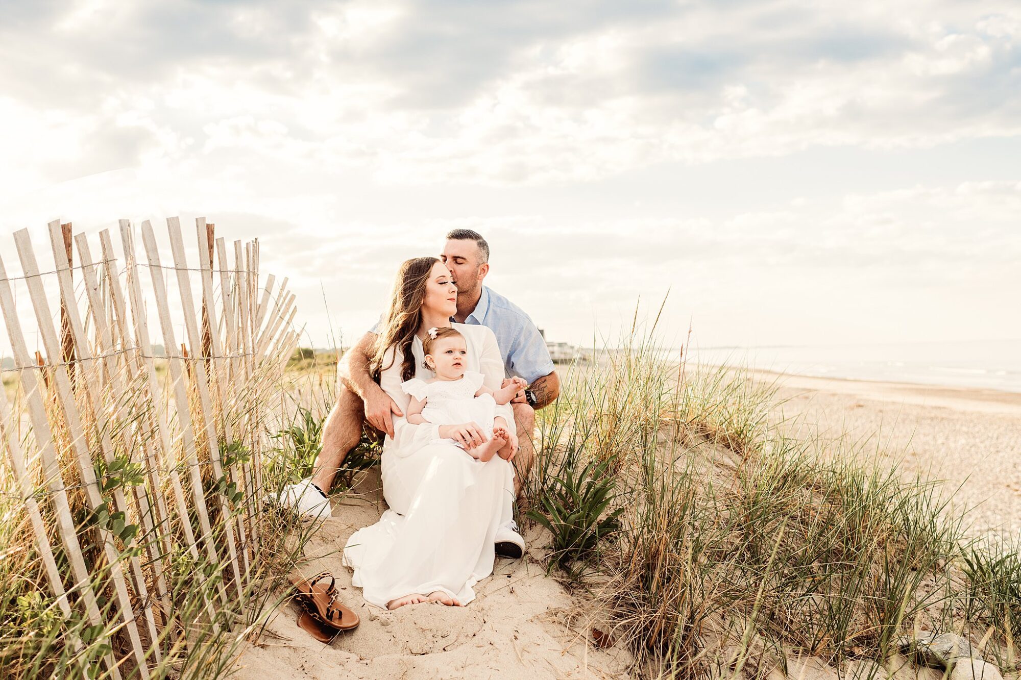 A family of three sits on sandy dunes by the beach during their Rexhame Beach Family Portraits. The mother holds a young child in her lap while the father sits behind, kissing her head, surrounded by beach grass and a bright, cloudy sky.