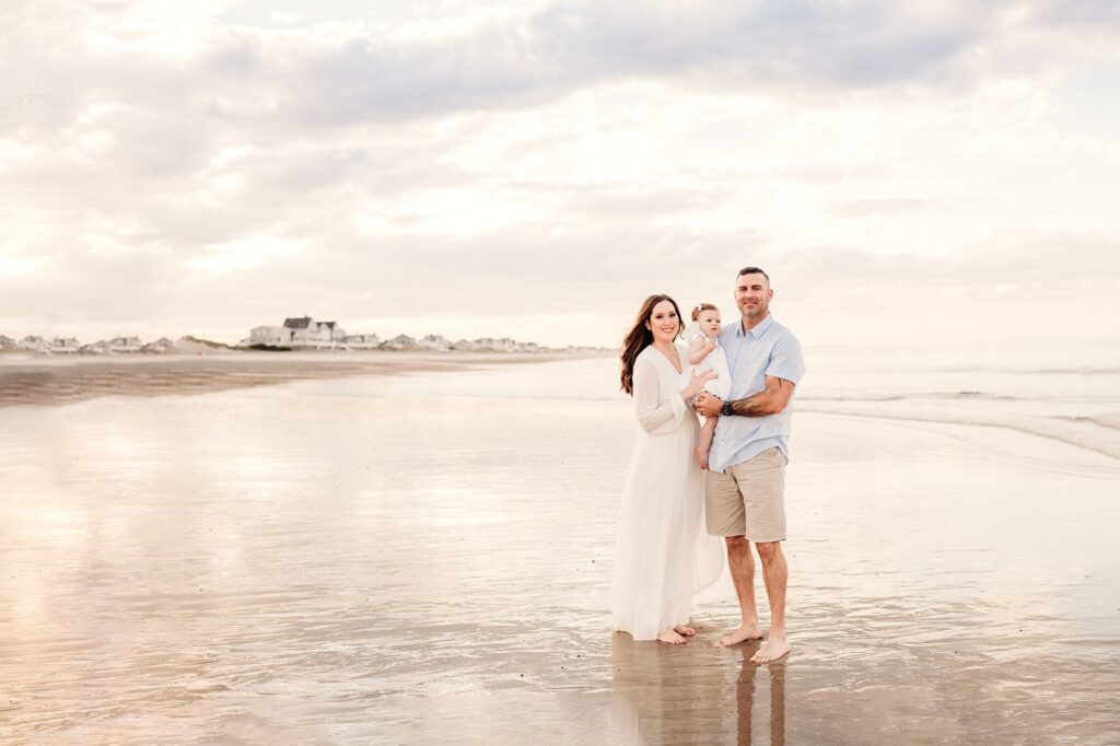 A smiling couple stands barefoot on Rexhame Beach with their baby under a cloudy sky. Dressed in light tones, they enjoy gentle waves and distant houses in the background—capturing timeless Family Portraits.