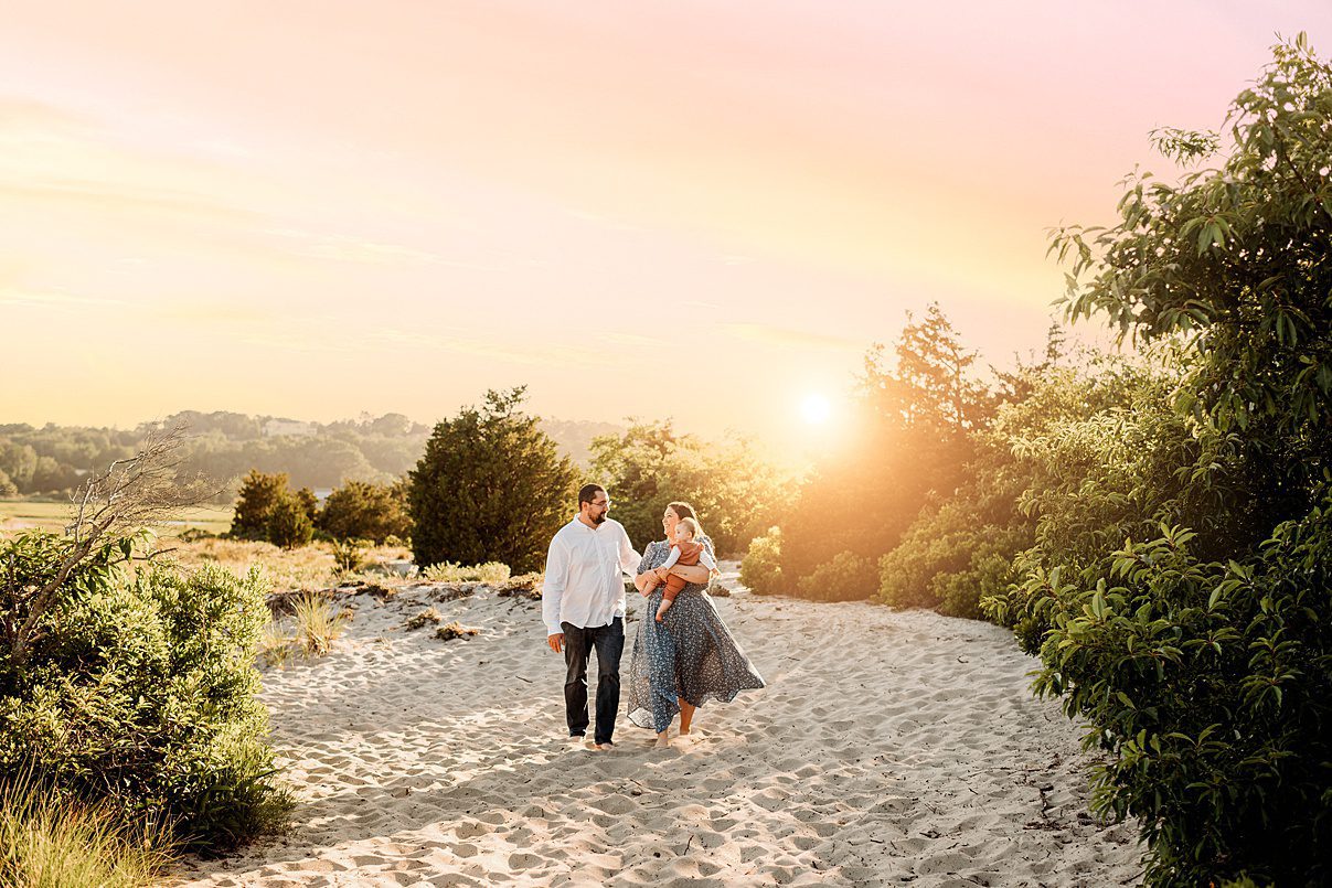 A couple walks barefoot on a sandy path at Rexhame Beach, surrounded by greenery and holding a baby, with a warm sunset glowing in the background—capturing the essence of family portraits.