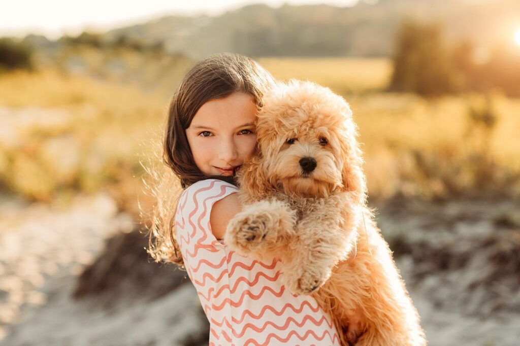 A young girl with long brown hair holds a fluffy, light brown puppy while standing outdoors in a sunlit field. Perfect for Rexhame Beach Family Portraits, both look towards the camera as golden light softly blurs the background.