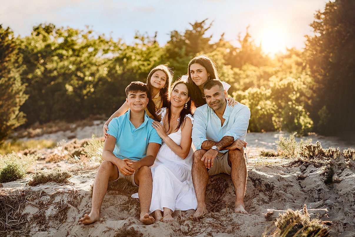 A family of five sits closely together on a sandy dune at sunset, surrounded by greenery. Smiling in light summer clothes, they create a warm, happy atmosphere—perfect for Rexhame Beach Family Portraits.