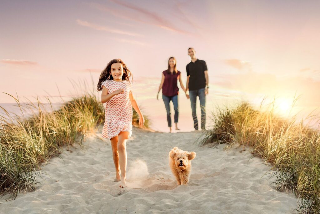 A young girl and a small dog run along a sandy path at sunset, two adults walking behind them. Grassy dunes frame the scene, making it perfect for memorable Rexhame Beach Family Portraits in the softly lit evening sky.