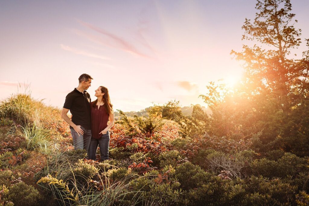 A couple stands together in a field of colorful bushes at sunset, smiling and looking at each other, with trees and a glowing sun in the background—capturing the warmth of Rexhame Beach Family Portraits.