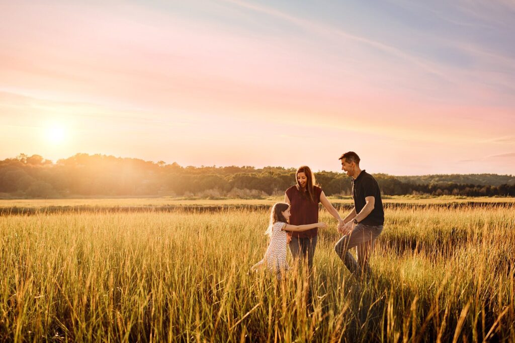 A family of three, two adults and a child, stand and play together in a sunlit field of tall grass at sunset, capturing the joy of Rexhame Beach Family Portraits with trees and a colorful sky in the background.