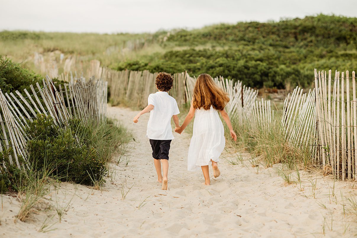 Two children, a boy and a girl, walk barefoot hand in hand down a sandy path between wooden fences and green dunes at Rexhame Beach, capturing the perfect setting for unforgettable family portraits on a sunny day.