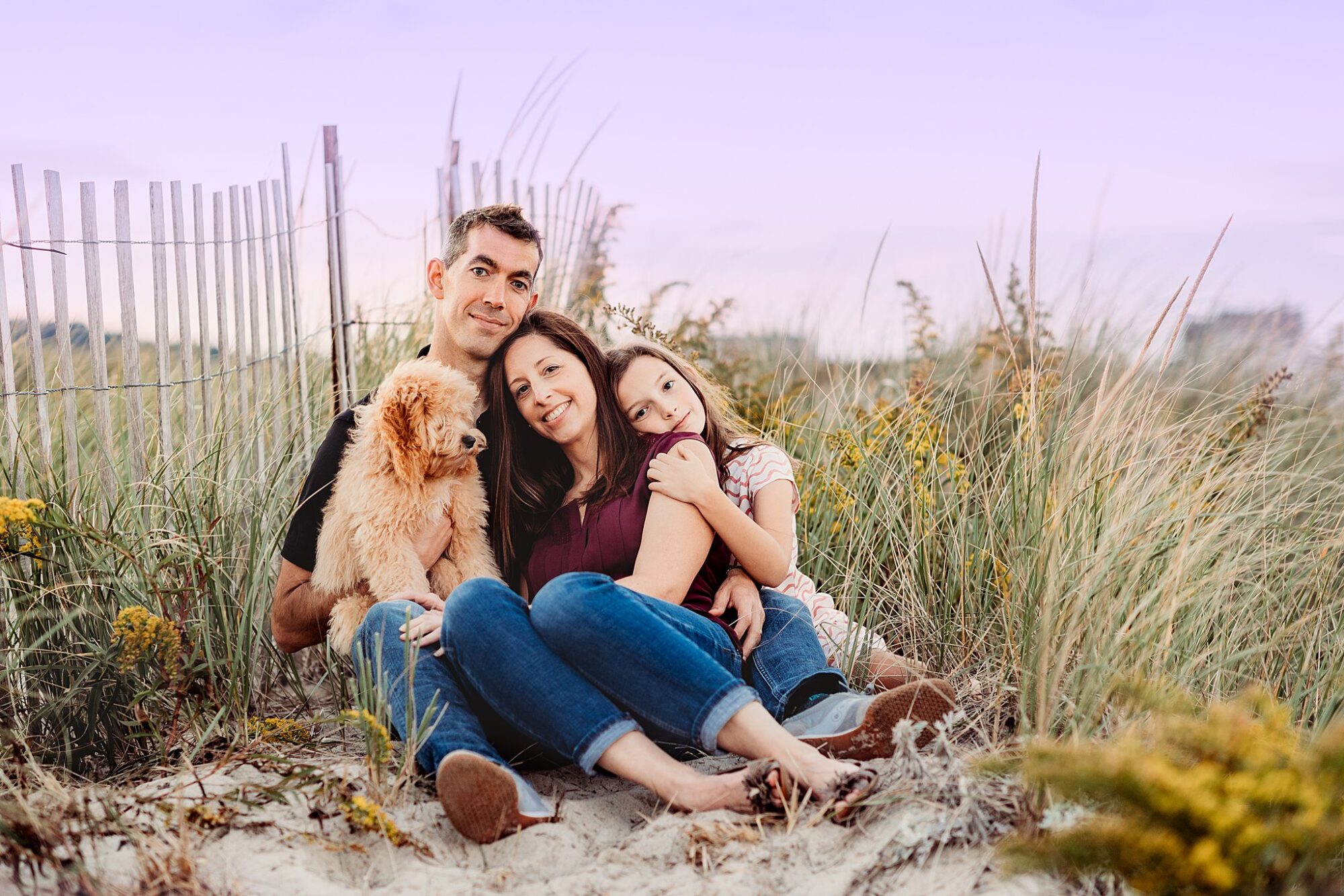 A family of three sits barefoot on sandy ground near beach grass and a wooden fence, cuddling a fluffy dog, all smiling softly under a pastel sky—capturing the warmth of Rexhame Beach Family Portraits.