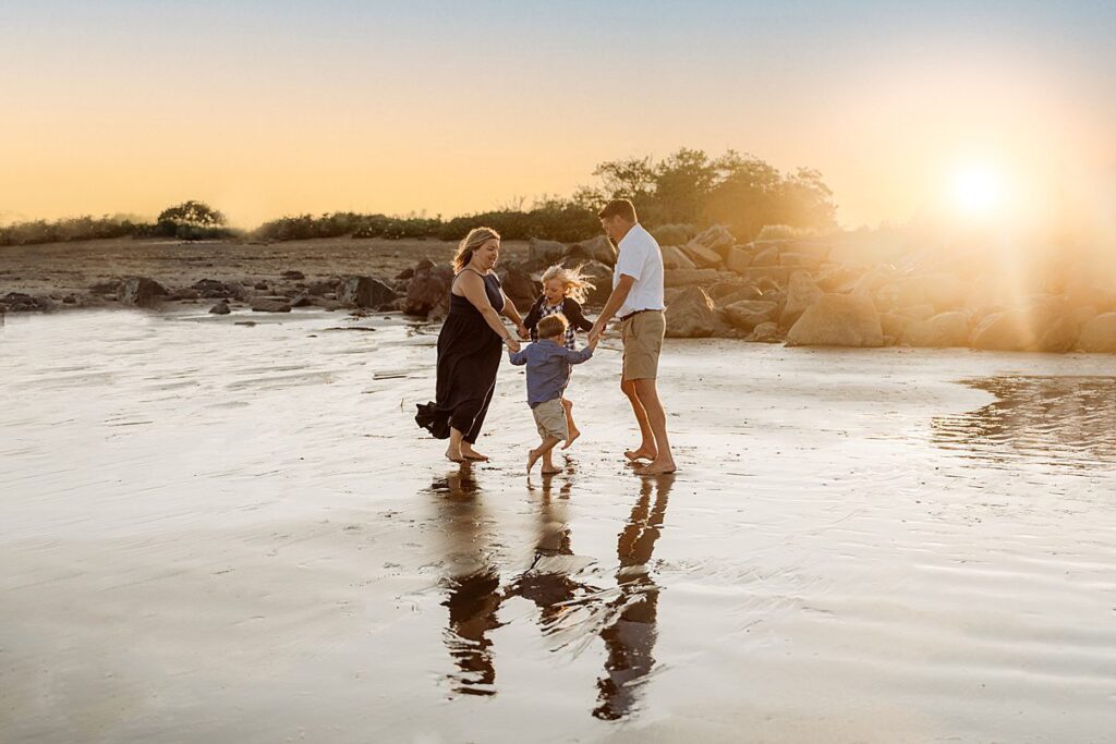 A family of three—two adults and a child—hold hands and spin together on a wet, reflective beach at sunset for their Rexhame Beach Family Portraits. Golden sky glows above as their reflections shimmer on the sand, with rocks and bushes in the background.