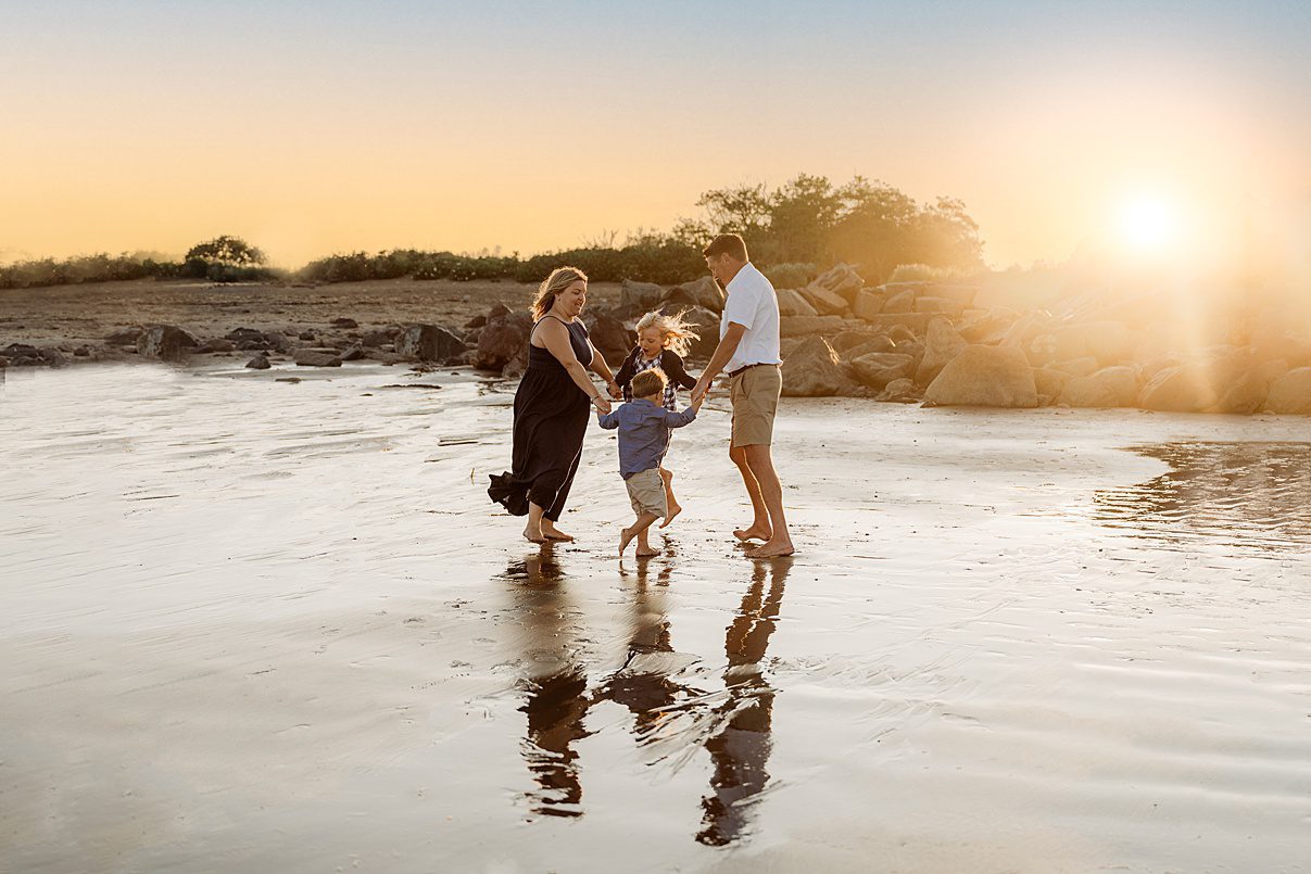 A family of three—two adults and a child—hold hands and spin together on a wet, reflective beach at sunset for their Rexhame Beach Family Portraits. Golden sky glows above as their reflections shimmer on the sand, with rocks and bushes in the background.