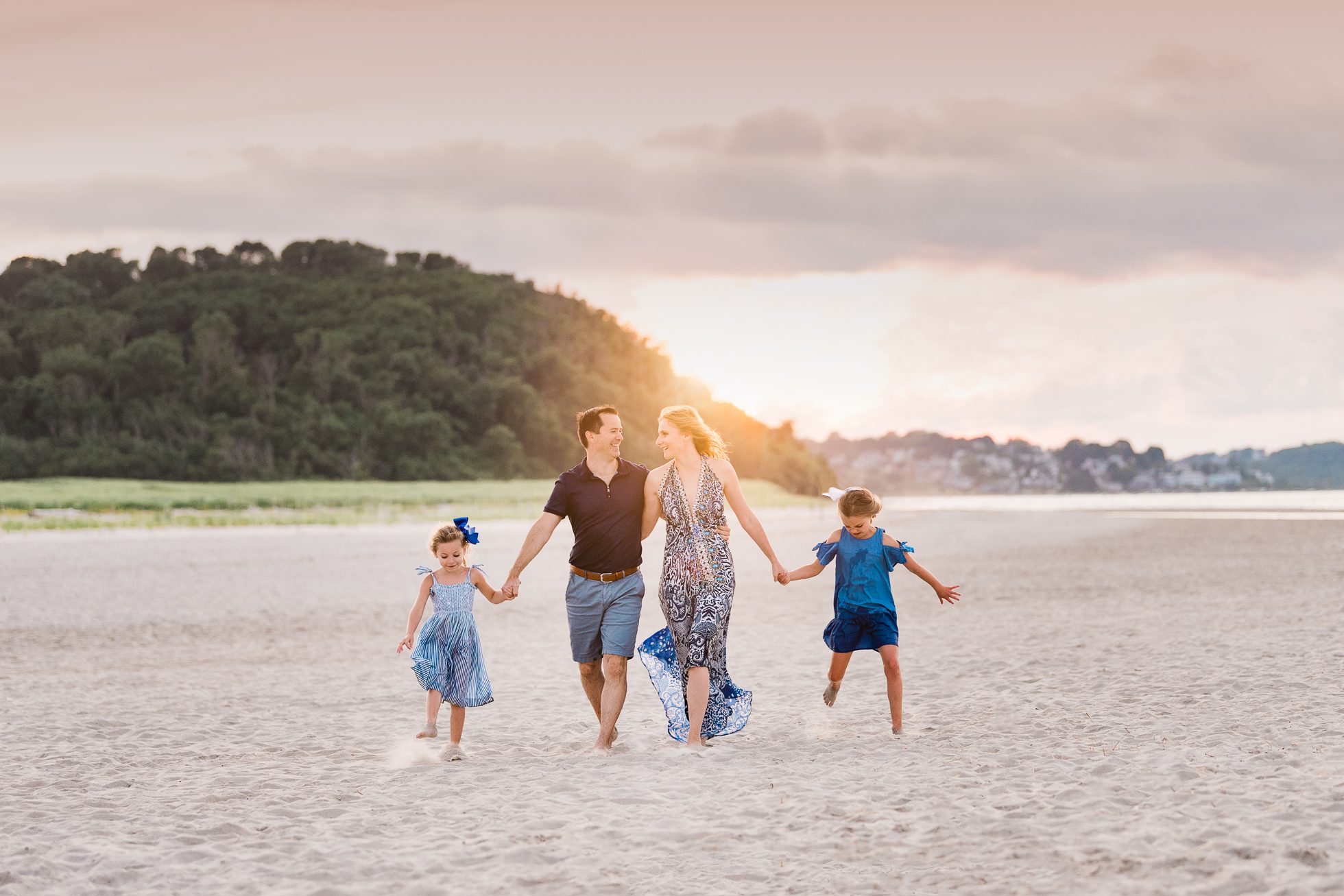 A family of four walks hand in hand on a sandy beach at sunset, greenery and distant buildings behind them. The parents smile as their daughters in blue dresses skip playfully—capturing the warmth of Rexhame Beach Family Portraits.