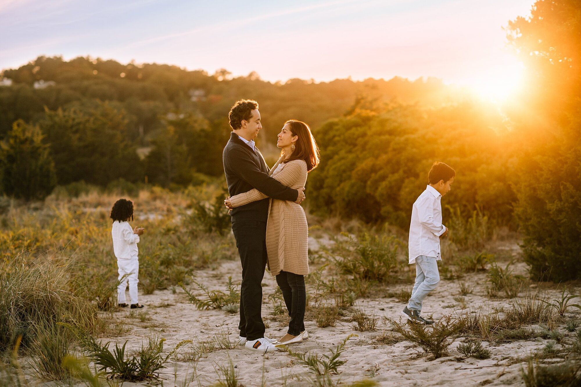 A couple embraces and smiles at each other on a sandy path at sunset during their Rexhame Beach Family Portraits, while two children explore the grassy area in the warm glow cast by the setting sun behind the trees.