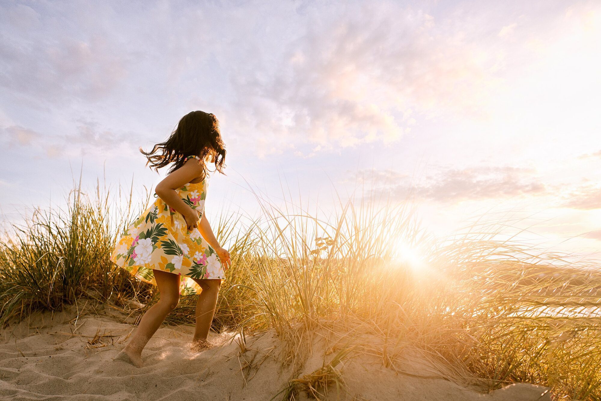 A young girl in a yellow floral dress runs barefoot up a sandy dune at Rexhame Beach, with tall grasses and a bright sunset illuminating the sky—perfect for family portraits.