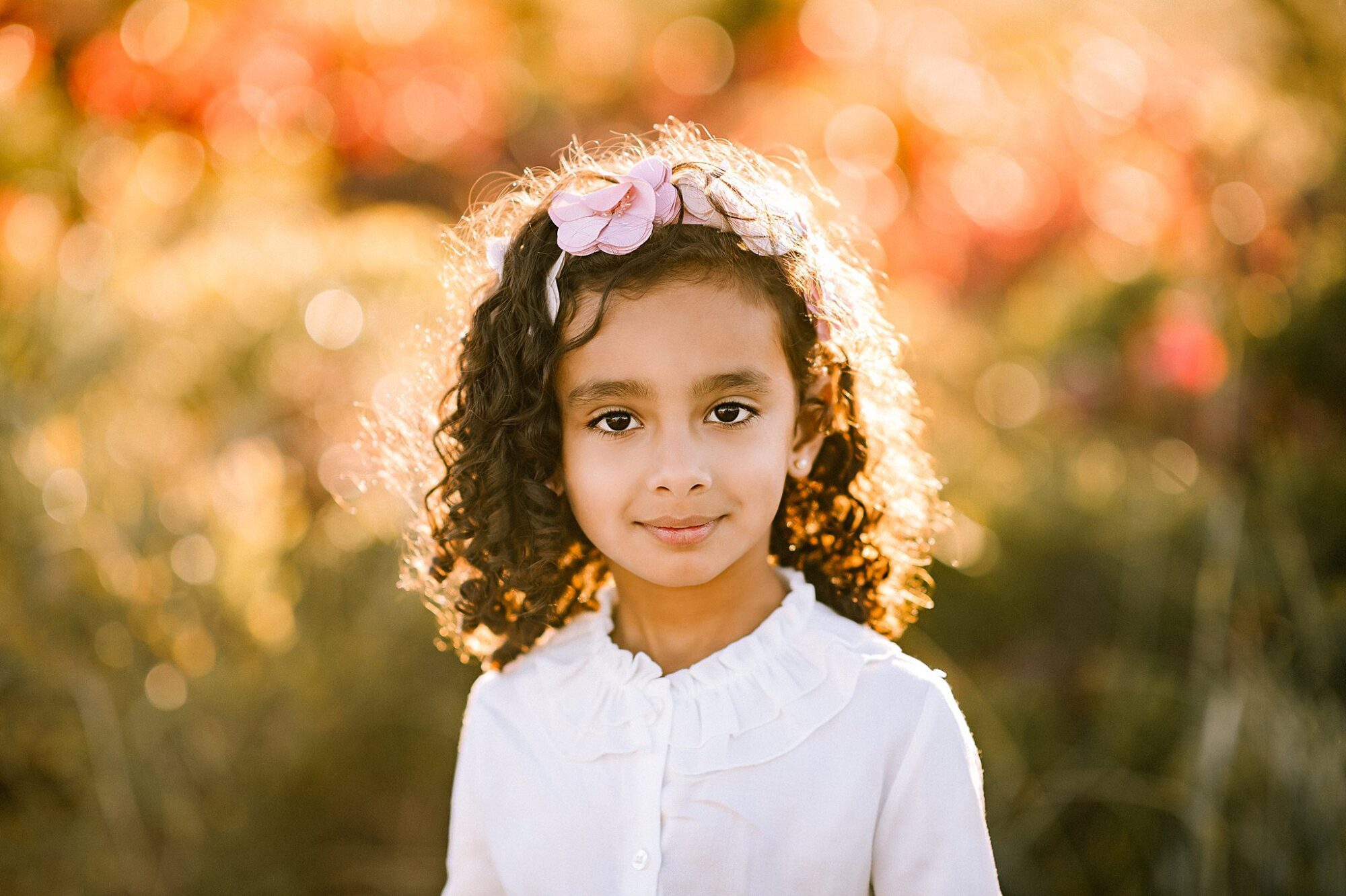 A young girl with curly hair and a pink flower headband stands outdoors in this Rexhame Beach Family Portrait, wearing a white ruffled blouse. The softly blurred background glows with warm, golden sunlight and bokeh effects.