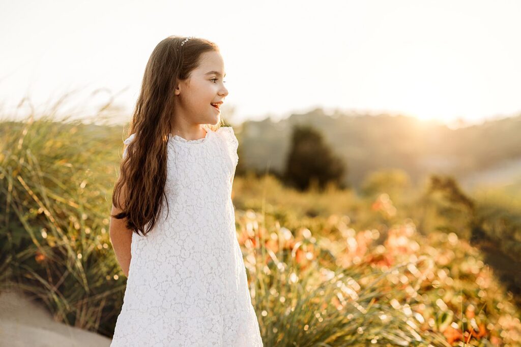 A young girl in a white lace dress stands outdoors, smiling and looking to the side. She has long brown hair, and the scene evokes Rexhame Beach Family Portraits with greenery and flowers bathed in warm, golden sunlight.