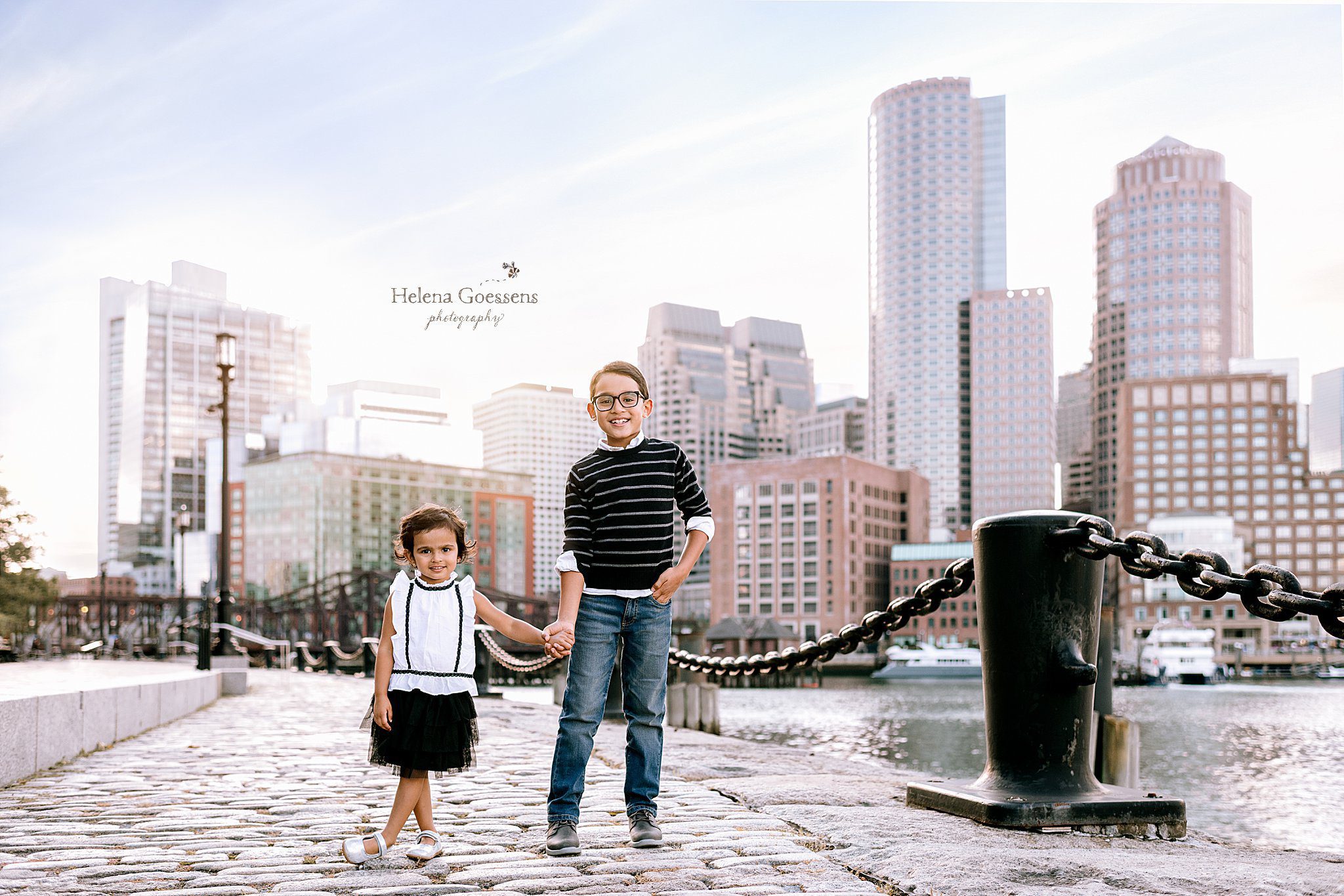 siblings hold hand standing in front of Boston skyline photographed by Helena Goessens Photography