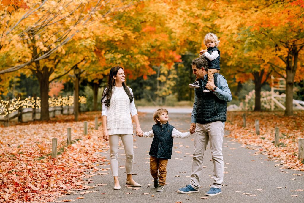 A family of four strolls along a leaf strewn path in an autumn park, vibrant trees painting the background. One child sits atop the man's shoulders, and another grasps both adults' hands—a picture perfect moment reminiscent of fall portraits at Bradley Estate.