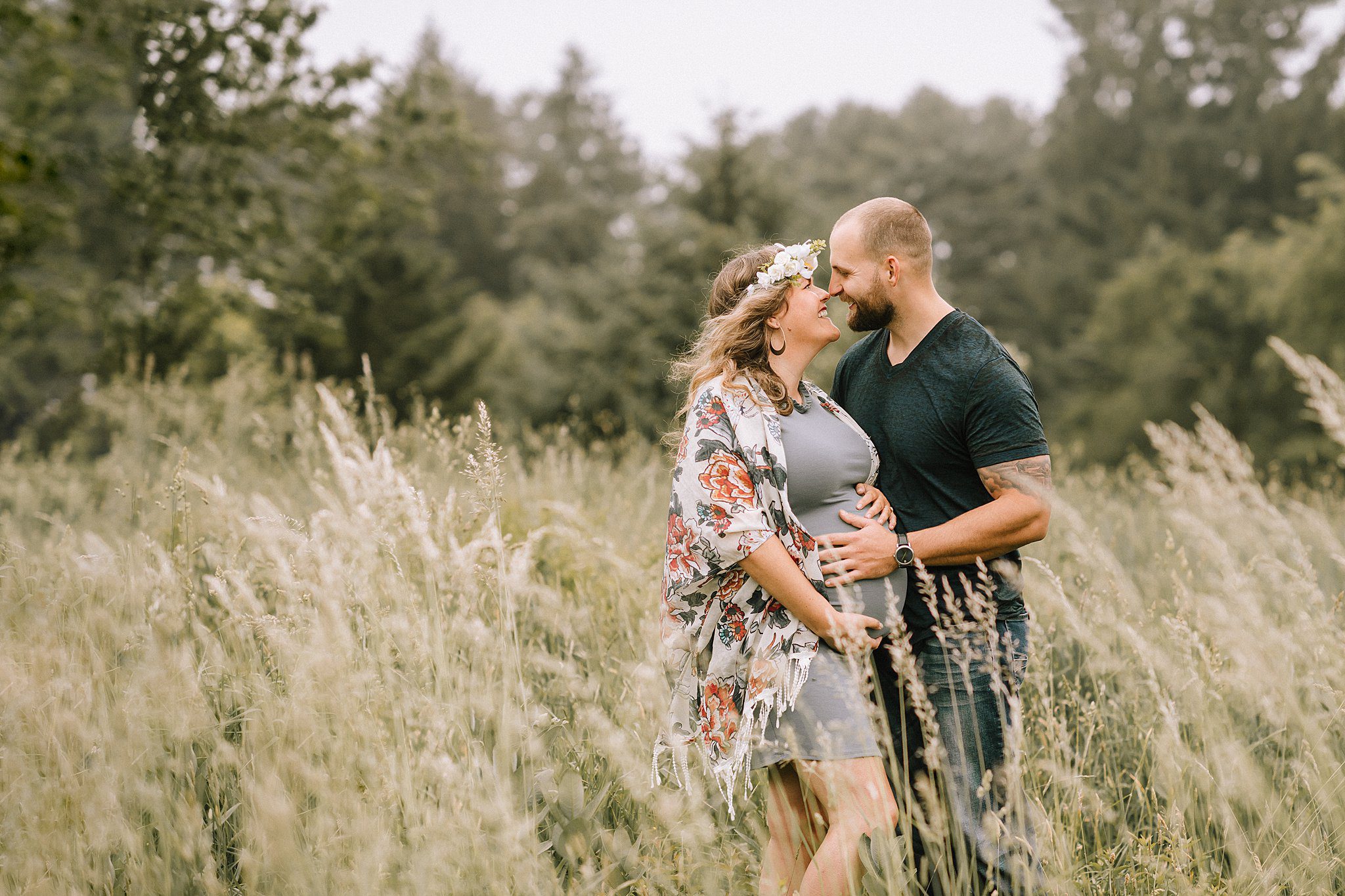 A couple stands in a lush field, surrounded by tall grass. The woman, embraced by the man in a dark shirt, wears a floral wrap and flower crown as she cradles her maternity bump. Both are smiling at each other, with a backdrop of trees.