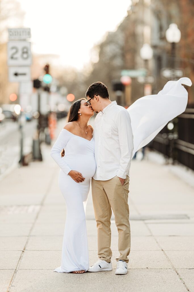 A pregnant woman in a flowing white maternity dress kisses a man in a white shirt and beige pants on a city sidewalk, tenderly holding his face with one hand. The elegance of her attire contrasts with the urban backdrop, marked by a speed limit sign visible in the distance.