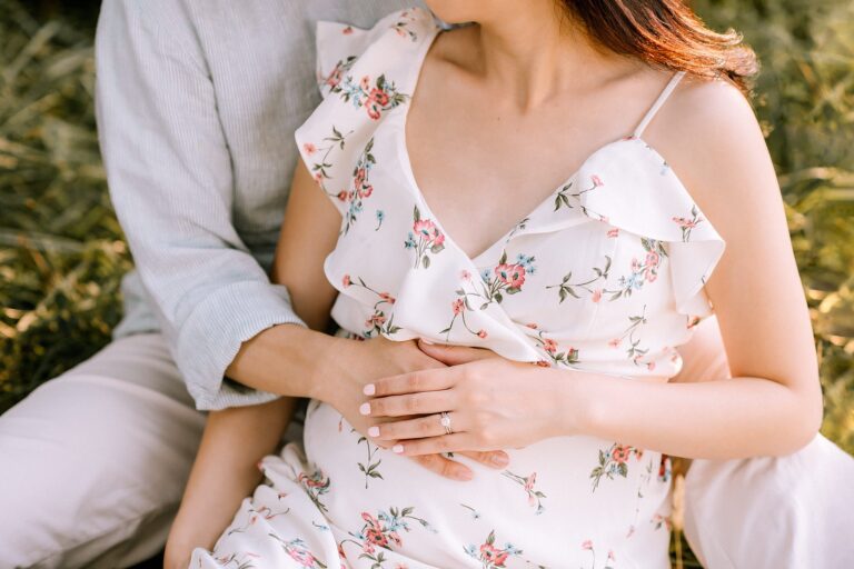 A couple sitting in a grassy area embraces tenderly, embodying the essence of maternity. The woman, in a floral dress, gently rests her hand over another on her pregnant belly, showcasing a ring. They sit close, radiating warmth and anticipation.