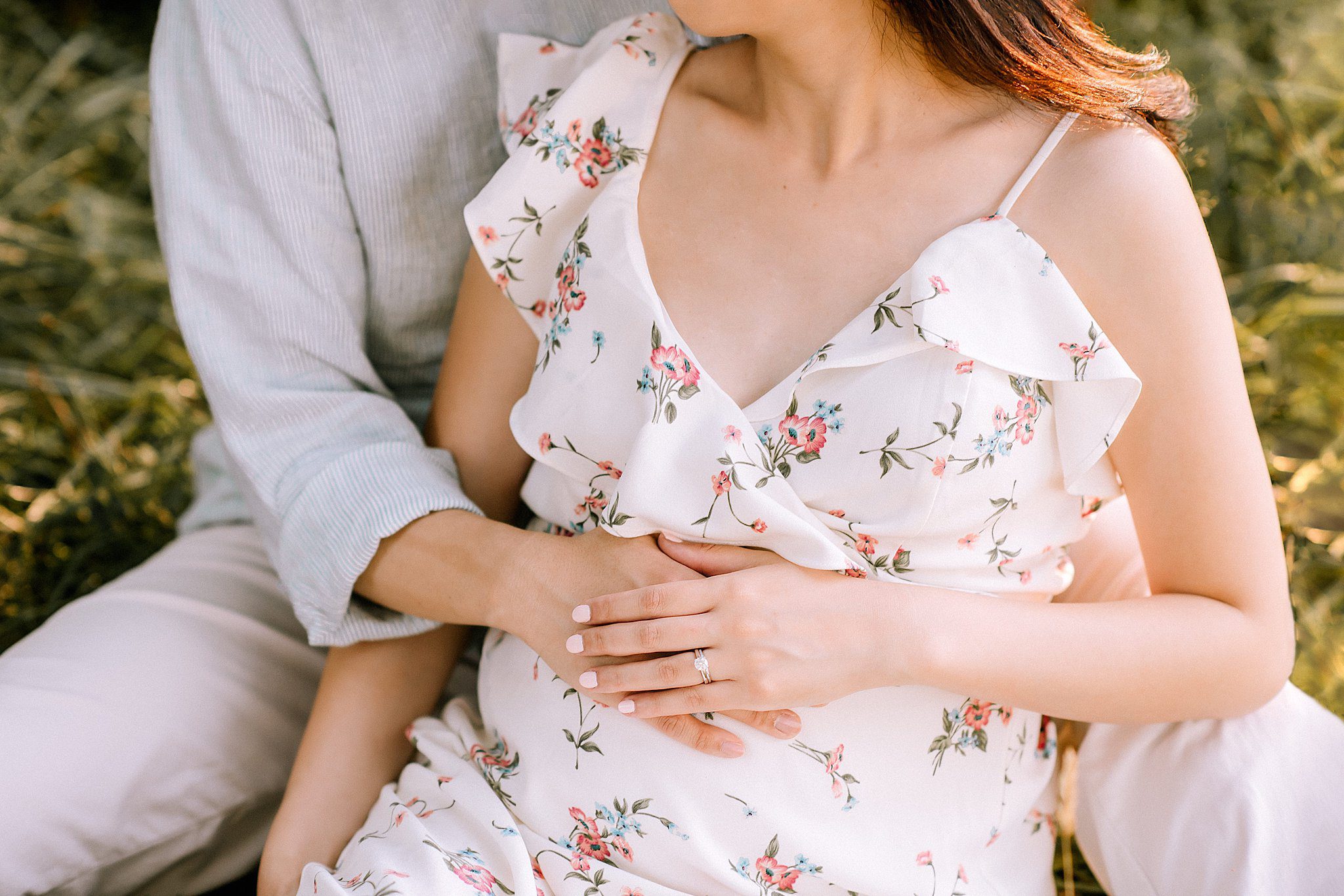 A couple sitting in a grassy area embraces tenderly, embodying the essence of maternity. The woman, in a floral dress, gently rests her hand over another on her pregnant belly, showcasing a ring. They sit close, radiating warmth and anticipation.
