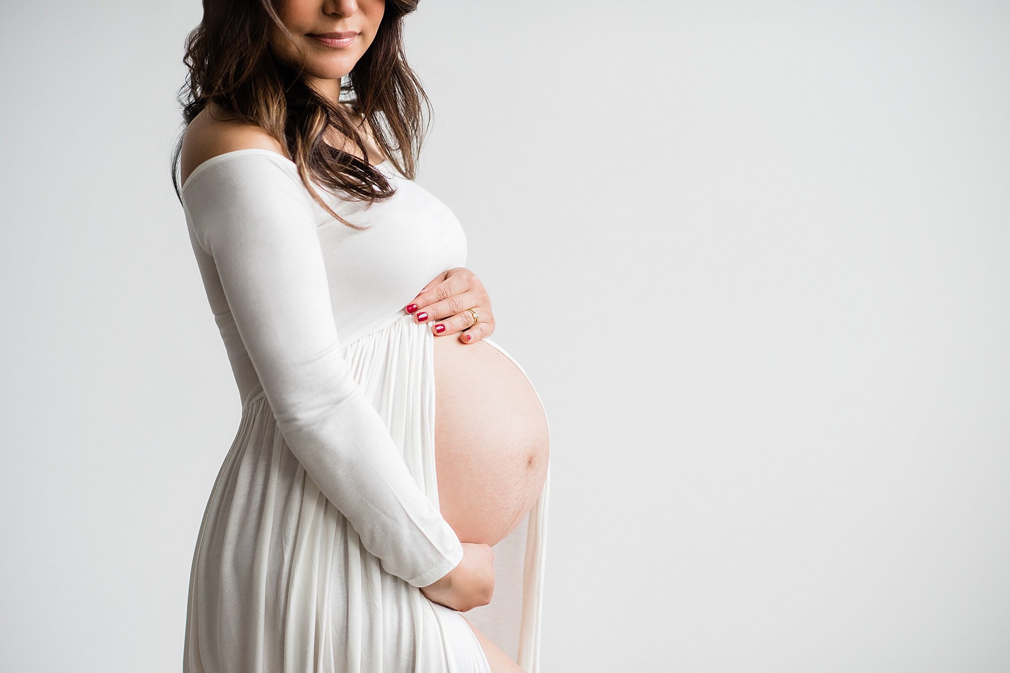 A pregnant woman, embodying maternity elegance, stands against a plain light background in a white off shoulder dress. Her long hair cascades over her shoulders as she gently holds her belly with red polished nails.