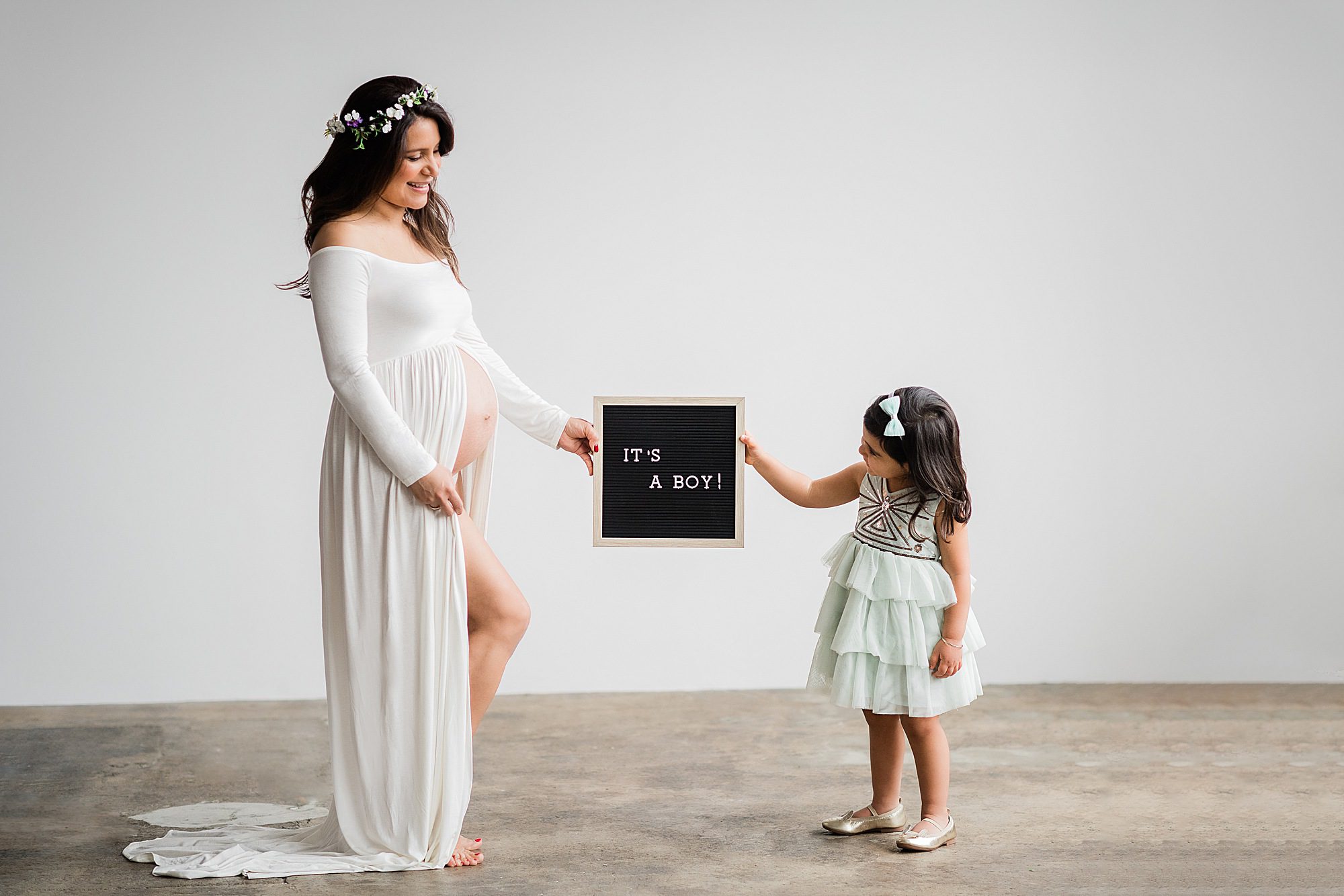 A pregnant woman in a flowing white dress holds a maternity letter board that reads “IT’S A BOY!” towards a young girl in a mint green dress. The girl reaches out to touch the board, both smiling in the light, minimalistic setting.