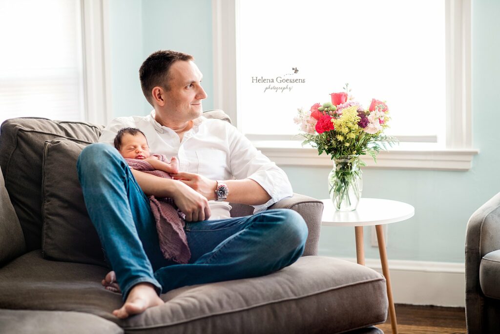dad holds baby girl on sofa during newborn photos at home with Boston newborn photographer  