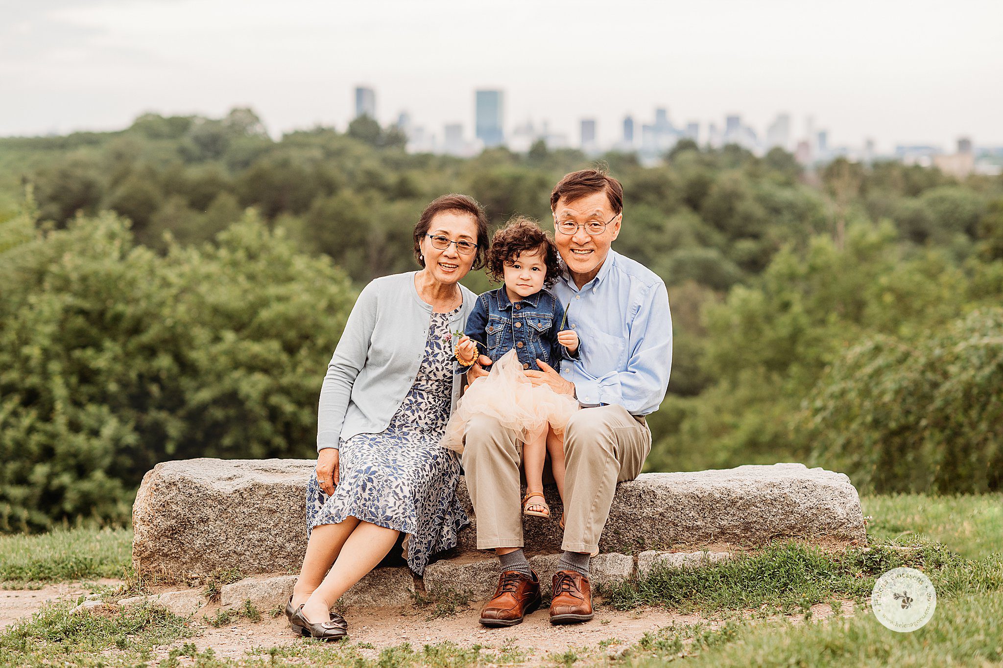 grandparents and toddler sit on rock with Boston in background