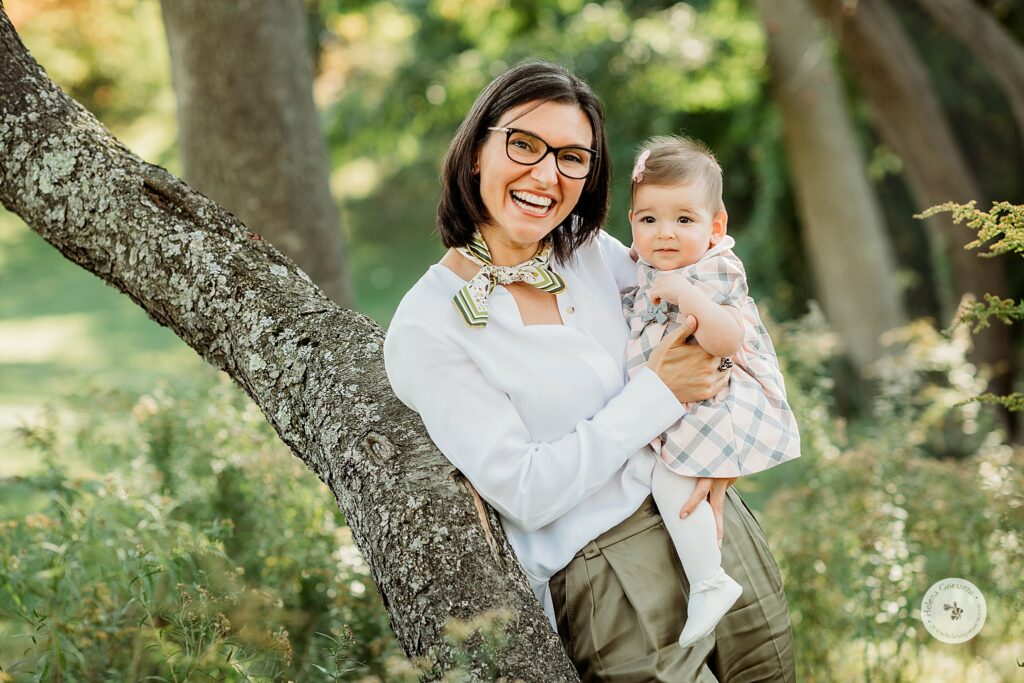 mom leans against tree holding baby girl during fall family photos in Brookline MA