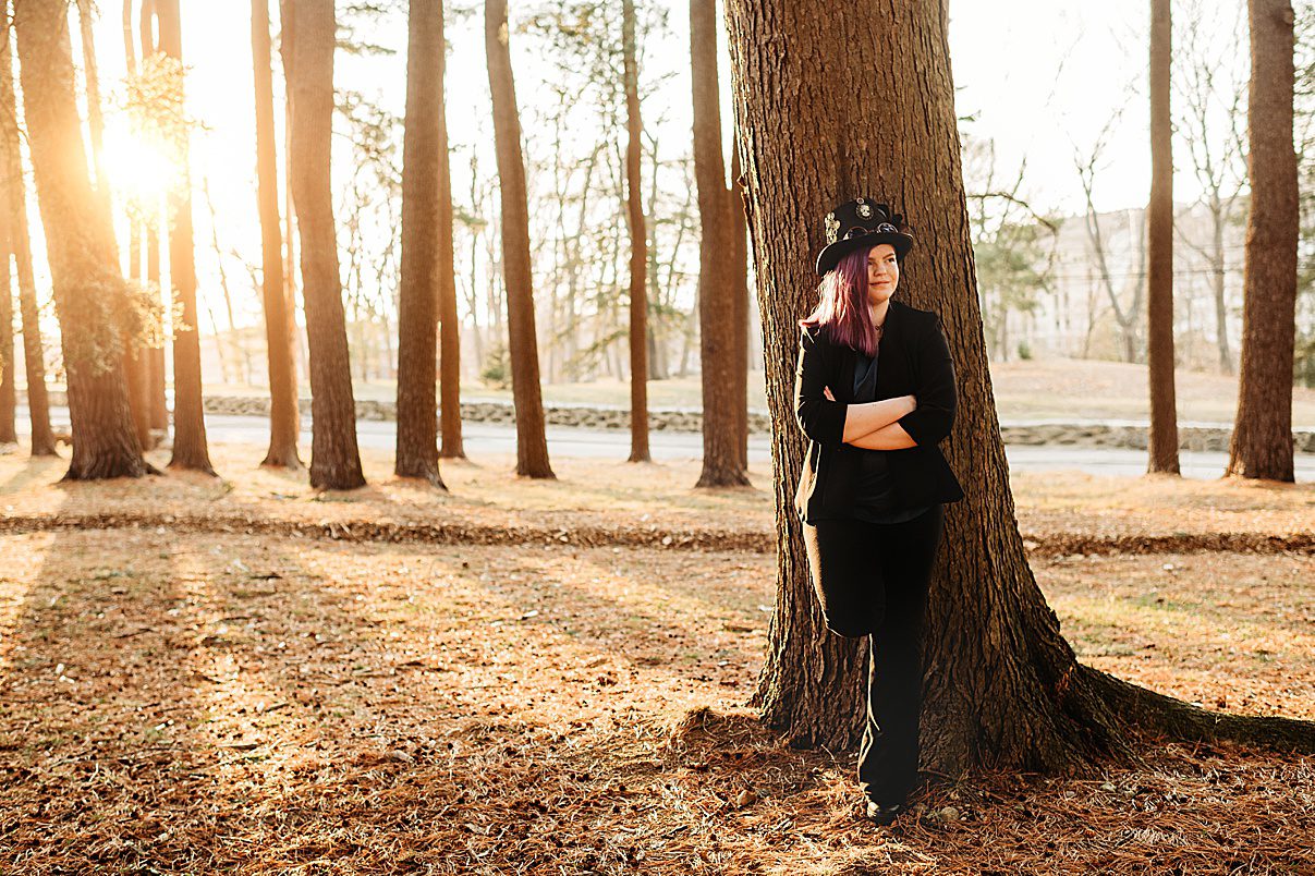 senior in custom hat leans against tree in Arnold's Arboretum during senior portraits 